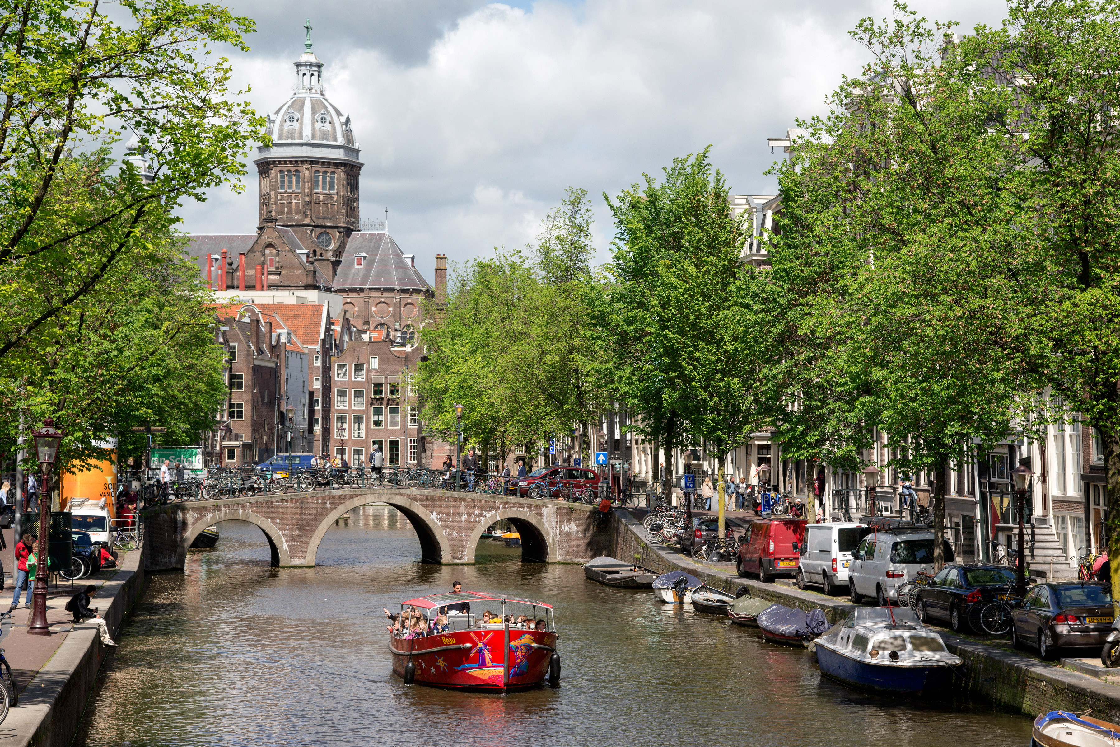 A red boat floating on a canal in Amsterdam, with cars lined on the side of the canal and a historcal building in the view
