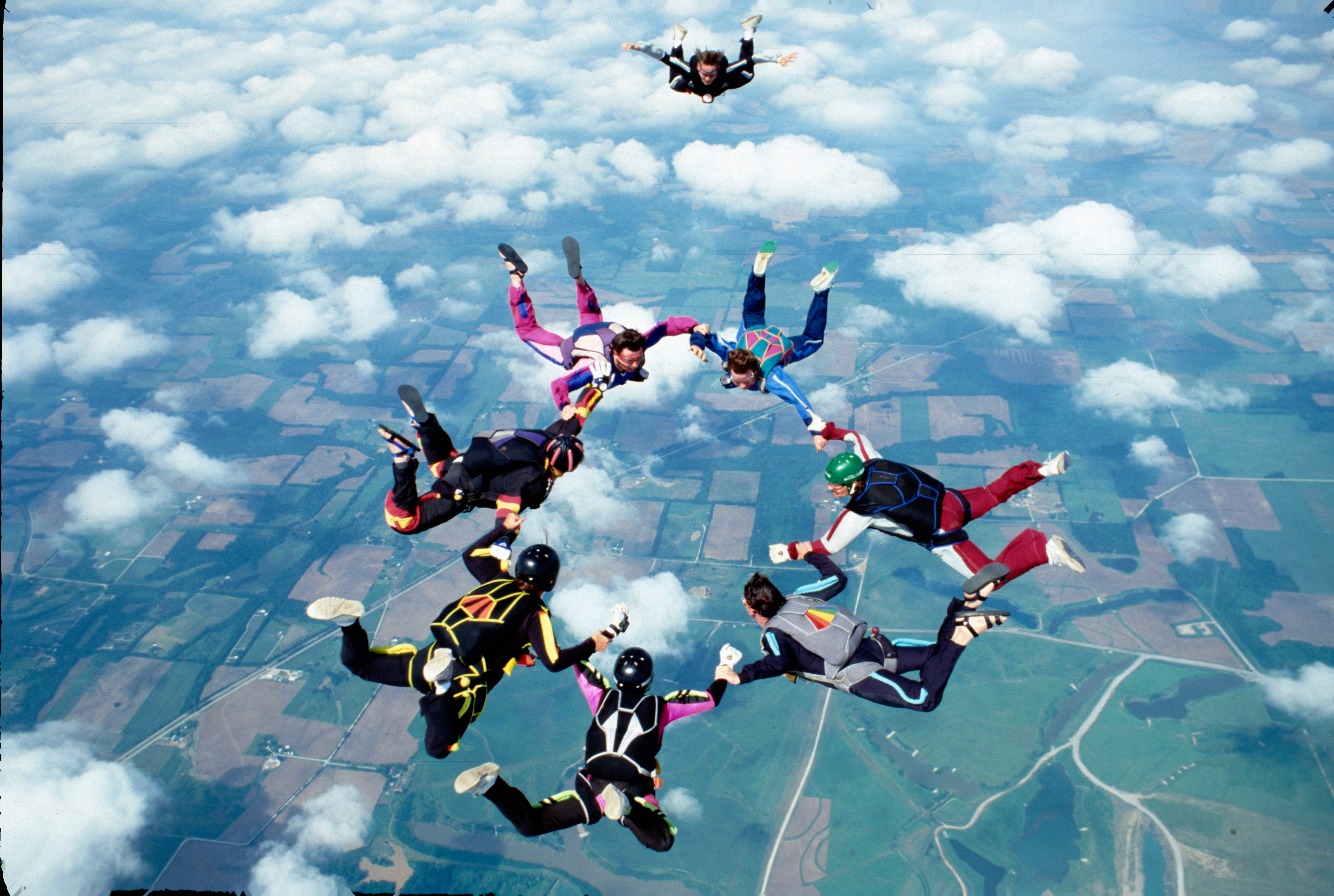 Seven people in a circle, holding hands as they skydive amongst the clouds and above green fields.