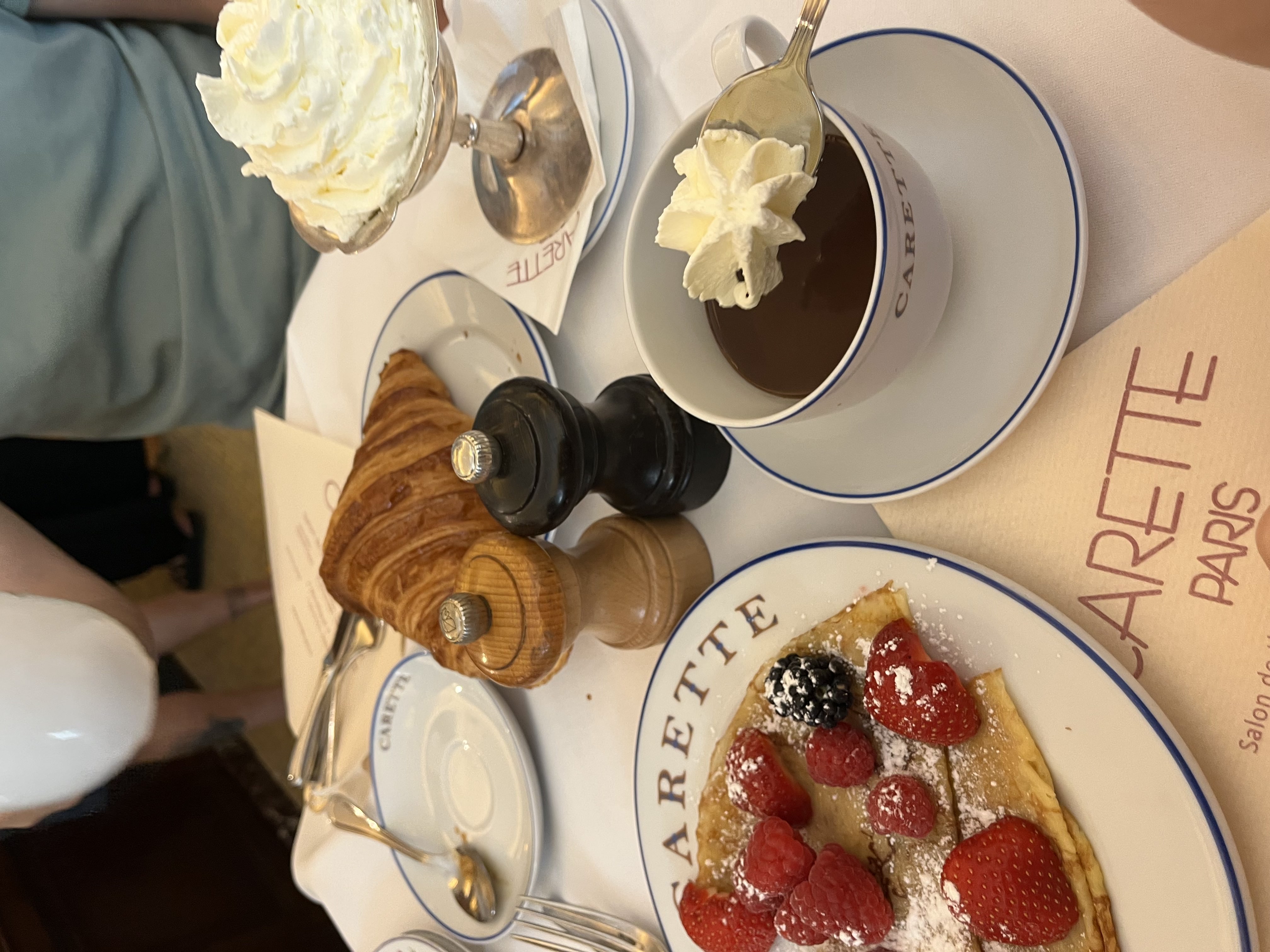 A picture of a croissant, hot chocolate with whipped cream, and a crepe with berries on a table at a cafe in paris.
